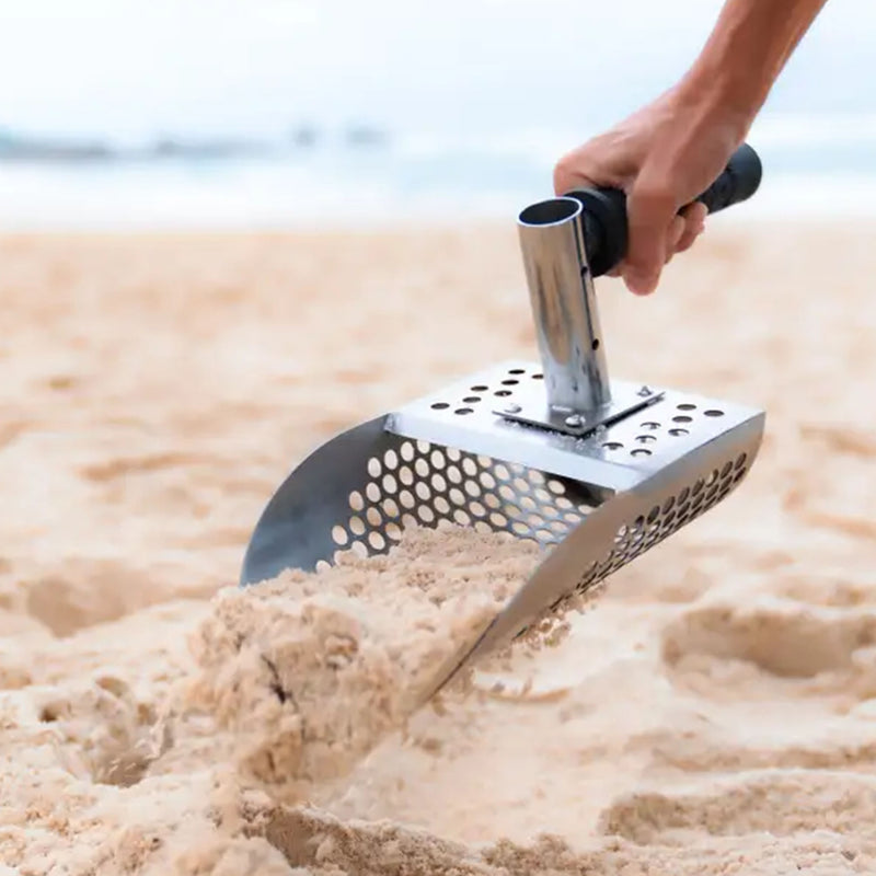 A stainless steel beach scoop with a perforated design and a black-handled grip being used on a sandy beach.