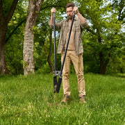 A man standing in a grassy park holding a long-handled black garden post hole digger with trees in the background.