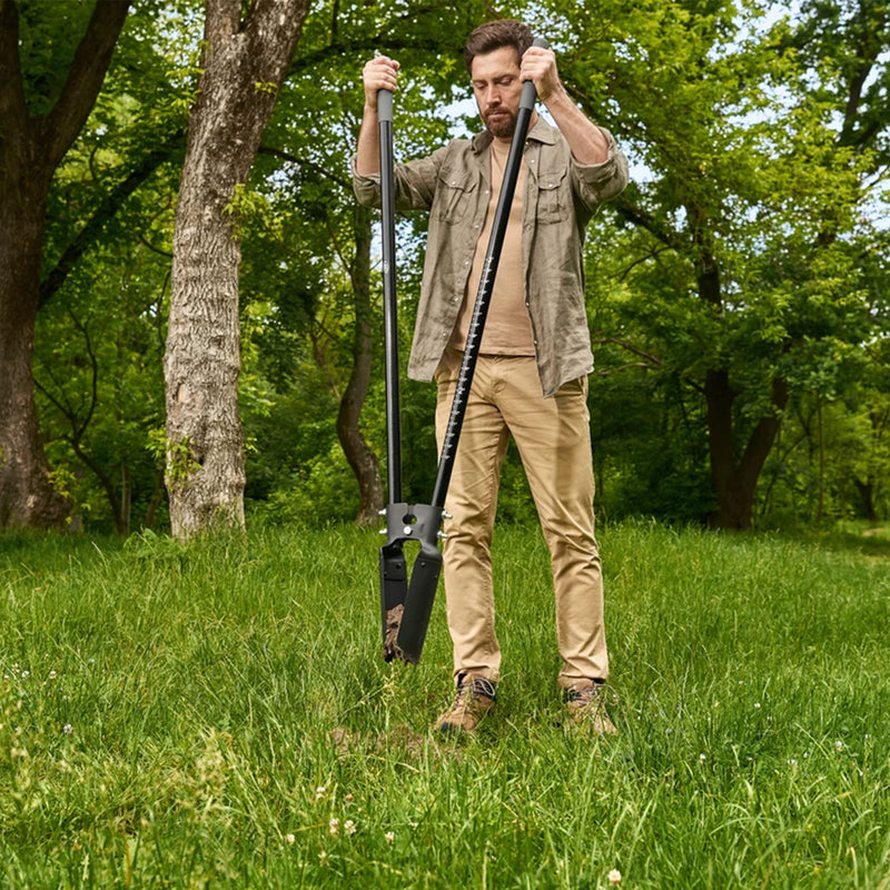 A man standing in a grassy park holding a long-handled black garden post hole digger with trees in the background.