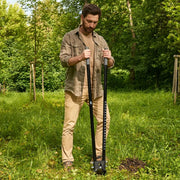A man standing in a grassy yard holding a black manual soil auger with a depth scale, surrounded by green trees and grass.