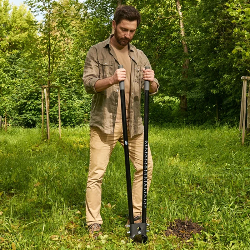 A man standing in a grassy yard holding a black manual soil auger with a depth scale, surrounded by green trees and grass.